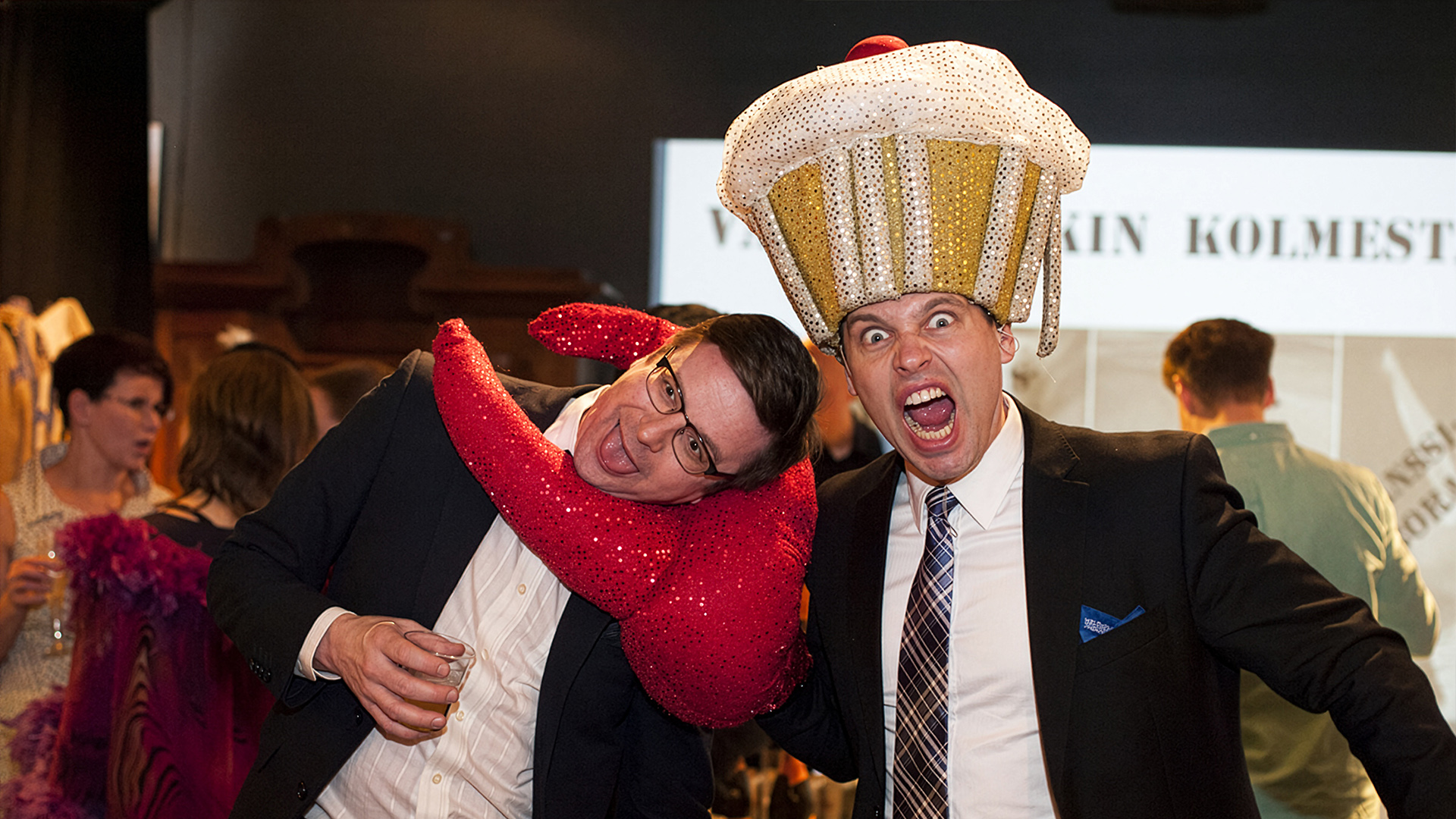 Muffinssihattuun pukeutunut, juhlapukuinen mies pitää toista miestä kaulasta kädessään olevalla jättimäisellä paljettikoristeisella ravunsaksella.