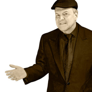 Taikuri Timo Kulmakko tummassa puvussa, oikea käsi ojennettuna kämmen ylöspäin.