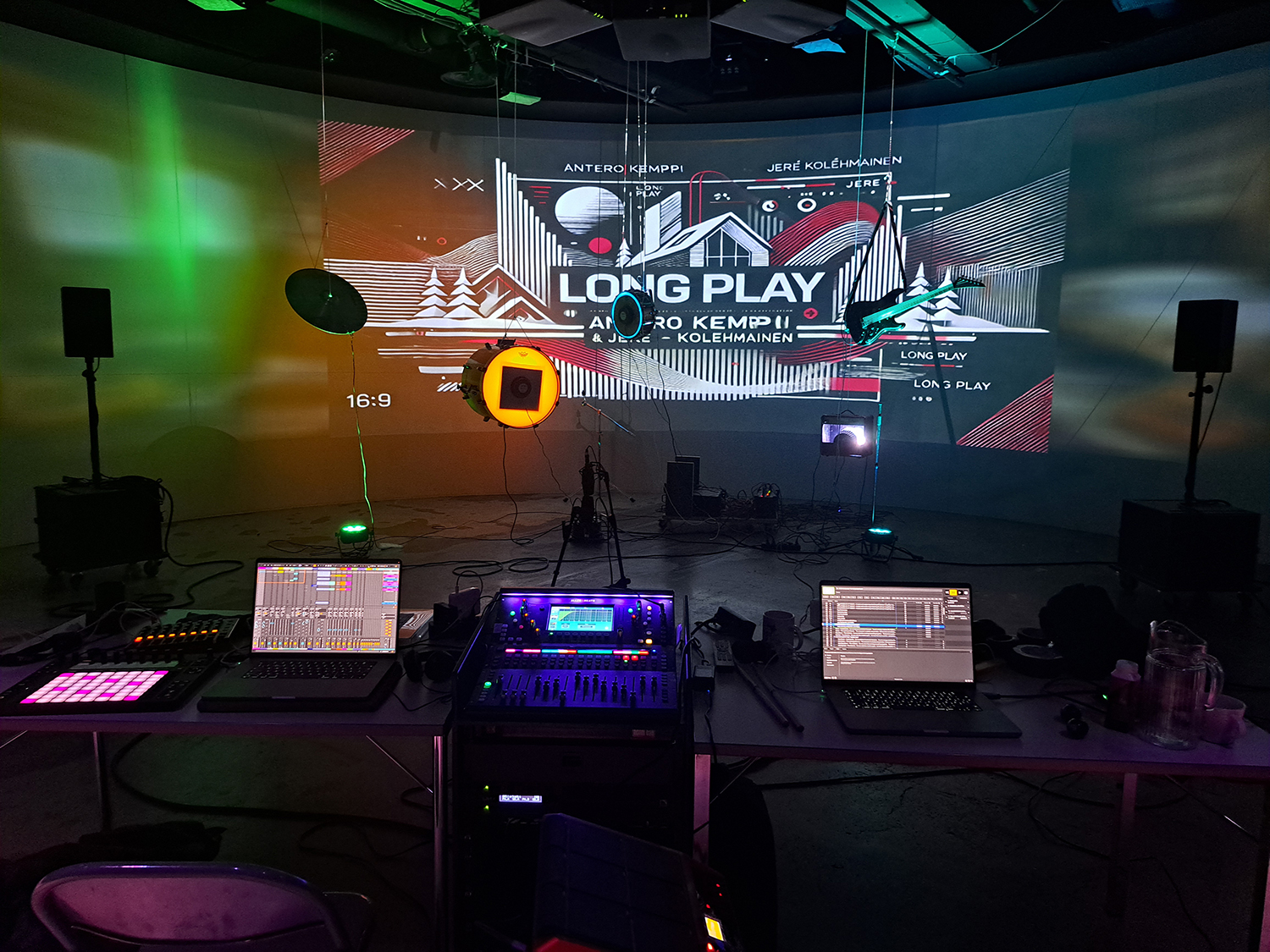 177 / 5 000 Musikinstrument upplysta med olika färgade lampor hänger från taket, inklusive en cymbal, en bastrumma, en mindre trumma och en elgitarr. I förgrunden syns ett ljudbord, ett ljusbord, en dator och annan utrustning.