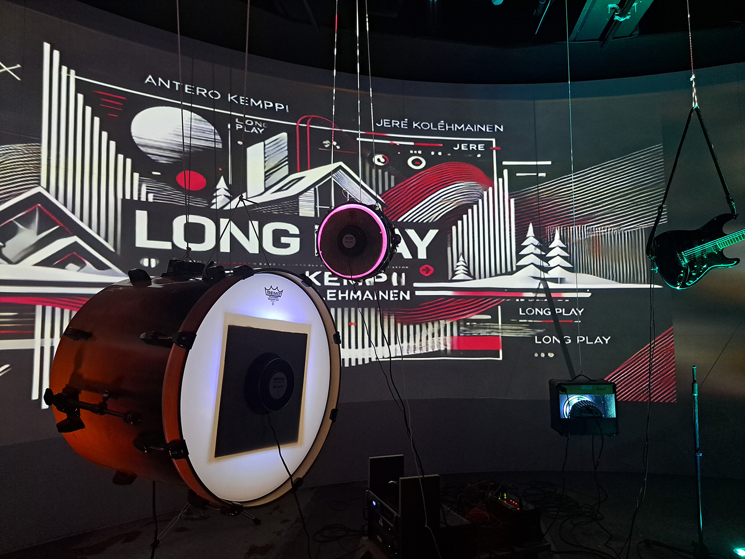 En bastrumma, en mindre trumma och en elgitarr hänger i luften. I bakgrunden projiceras texten Long Play på väggen.